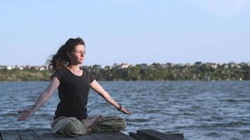 Seitenansicht einer schlanken jungen Frau, die im Lotussitz auf einem Pier sitzt und ihre Hände hebt. Frau, die Yoga am Strand bei Sonnenuntergang gegen das Wasser praktiziert. 4k-Zeitlupenvideo. video