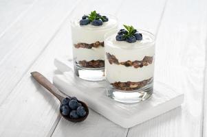 postre con requesón, arándanos frescos y granola en un vaso sobre un fondo blanco de madera. foto