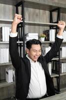 Employee employer employment entrepreneur executive man in formalwear raised hands fists up sit on chair. Emotional guy feeling glad and happy as he's great success, finishes successful business. photo