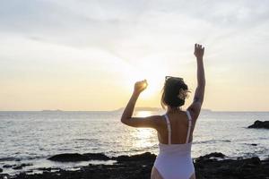 mujer turista levantando la mano con puesta de sol en la playa en viaje de vacaciones. mujer con traje de baño relajante y brazos levantados con granos de arena en la piel. dama asiática disfrutando de la naturaleza en vacaciones. concepto de libertad foto