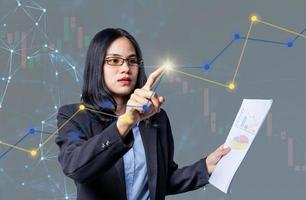 Confident businesswoman pressing a modern button on digital screen and holding bar graph sheet for trading against stock market and candlestick graphic on background. Woman analyzed stock graph chart photo
