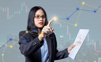 Confident businesswoman pressing a modern button on digital screen and holding bar graph sheet for trading against stock market and candlestick graphic on background. Woman analyzed stock graph chart photo