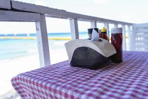 Close up of tissue box, sauce bottle, and seasoning on the table in restaurant beach in the morning at sunlight. Beach background photo