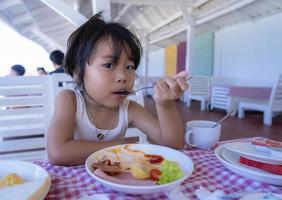 niña pequeña y linda desayunando sosteniendo una cuchara y manchando la comida alrededor de su boca y mirando la cámara. mujercita hambrienta a la hora del desayuno en el restaurante. niño y concepto de alimentación saludable. foto