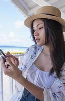 Young woman using smartphone by seaside. Happy tourist relaxing and using mobile phone for social network on beach of tropical sunrise uses communication technology. Travel concepts and technology photo