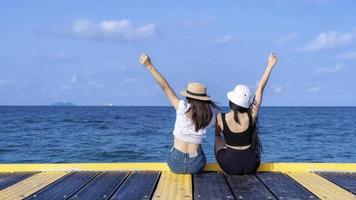 Back view of two happy young women rising hands up and sitting on  bridge with tropical ocean backgrounds, enjoying beautiful view of blue sea. Outdoor summer lifestyle. Vacations and freedom concept photo