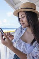 Young woman using smartphone by seaside. Happy tourist relaxing and using mobile phone for social network on beach of tropical sunrise uses communication technology. Travel concepts and technology photo