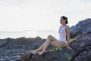 Portrait of beautiful fashionable woman in white bikini posing at the rock beach. Young Asian woman dressed in swimsuit laying on a rock on a summer day with sunset backgrounds at Koh Larn in Pattaya photo