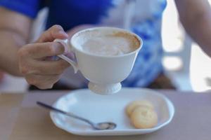 Close up of lady's hands holding white coffee cup with blur cookies at cafe table during coffee break or breakfast in vacations. Traveler woman holding white mug of hot coffee for relaxing on holidays photo