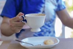 Close up of lady's hands holding white coffee cup with blur cookies at cafe table during coffee break or breakfast in vacations. Traveler woman holding white mug of hot coffee for relaxing on holidays photo