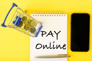 PAY online, the text is written in a notebook lying on a yellow background and a shopping cart with coins one Euro, one dollar. Flat lay. photo