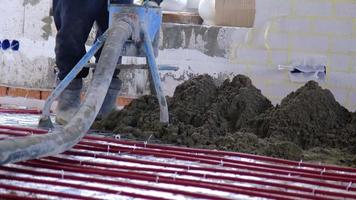 solado de suelo semiseco: un trabajador palea una mezcla de construcción a través de un manguito especial para cementar y nivelar tuberías de calefacción por suelo radiante. camara lenta video