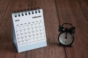 February 2023 white desk calendar on wooden table with alarm clock pointing at 12 o'clock. photo