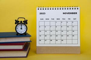 calendario de escritorio de noviembre de 2023 con libros y despertador sobre fondo amarillo. foto