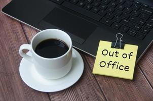 Coffee cup and laptop on wooden table with yellow notepad with text - Out of office. photo
