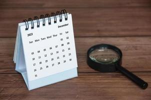 December 2023 white desk calendar with magnifying glass on wooden table. Calendar copy space photo