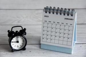 enero de 2023 calendario de escritorio blanco con despertador negro apuntando a las 9 en punto sobre una mesa de madera. foto