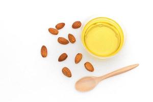 Clear yellow almond oil in a clear bowl with a wooden spoon on top of it and several almonds on a white background.top view, flat lay, top-down. photo