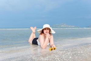 A plump white woman in a bathing suit or swimsuit, white hat, and yellow sunglasses is lying on the beach smiling prone. photo
