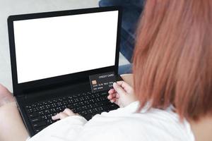 una mujer que tiene una tarjeta de crédito, que paga en línea, en una computadora portátil, sentada en el sofá de la sala de estar, en casa con un camino de recorte. el concepto de compras en línea. cuaderno de pantalla blanca para maqueta foto