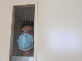 hombre solitario con mascarilla médica mirando por la ventana. aislamiento en casa para la autocuarentena. concepto de cuarentena domiciliaria, prevención covid-19. situación de brote de coronavirus foto