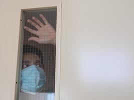 hombre solitario con mascarilla médica mirando por la ventana. aislamiento en casa para la autocuarentena. concepto de cuarentena domiciliaria, prevención covid-19. situación de brote de coronavirus foto