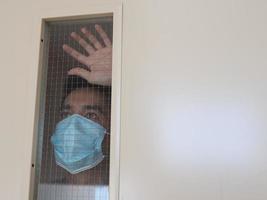 hombre solitario con mascarilla médica mirando por la ventana. aislamiento en casa para la autocuarentena. concepto de cuarentena domiciliaria, prevención covid-19. situación de brote de coronavirus foto