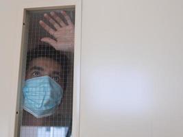 Lonely man in medical mask looking through the window. Isolation at home for self quarantine. Concept home quarantine, prevention COVID-19. Coronavirus outbreak situation photo