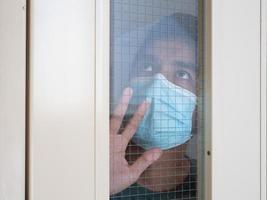Lonely man in medical mask looking through the window. Isolation at home for self quarantine. Concept home quarantine, prevention COVID-19. Coronavirus outbreak situation photo
