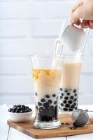 Making bubble tea, pouring blend milk tea into brown sugar pattern drinking glass cup on white wooden table background, close up, copy space photo