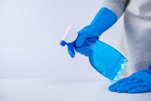 una joven ama de llaves en delantal está limpiando, limpiando la superficie de la mesa con guantes azules, trapo amarillo húmedo, limpiador de botellas rociadas, concepto de diseño cerrado. foto