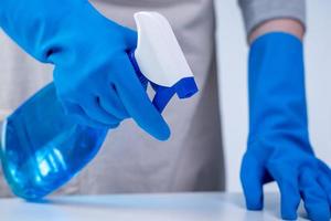 una joven ama de llaves en delantal está limpiando, limpiando la superficie de la mesa con guantes azules, trapo amarillo húmedo, limpiador de botellas rociadas, concepto de diseño cerrado. foto