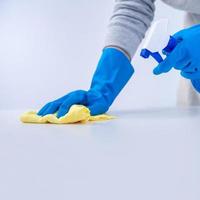 una joven ama de llaves en delantal está limpiando, limpiando la superficie de la mesa con guantes azules, trapo amarillo húmedo, limpiador de botellas rociadas, concepto de diseño cerrado. foto