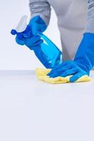 Young woman housekeeper in apron is cleaning, wiping down table surface with blue gloves, wet yellow rag, spraying bottle cleaner, closeup design concept. photo