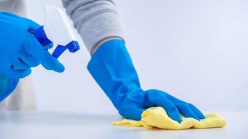 Young woman housekeeper in apron is cleaning, wiping down table surface with blue gloves, wet yellow rag, spraying bottle cleaner, closeup design concept. photo