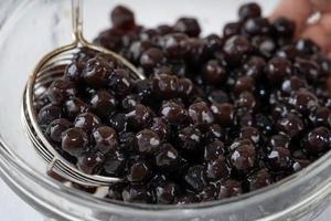 hacer té de burbujas, sacar y verter bolas de burbujas de perlas de tapioca con sabor a azúcar moreno cocido en una taza sobre fondo de mesa de madera blanca, cerrar, copiar espacio. foto