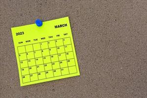 March 2023 yellow sticky note calendar with pin on cork bulletin billboard photo