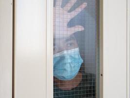 hombre solitario con mascarilla médica mirando por la ventana. aislamiento en casa para la autocuarentena. concepto de cuarentena domiciliaria, prevención covid-19. situación de brote de coronavirus foto