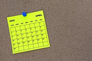 April 2023 yellow sticky note calendar with pin on cork bulletin billboard. photo