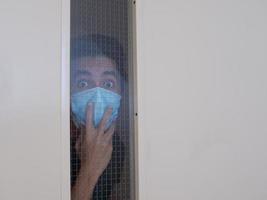 Lonely man in medical mask looking through the window. Isolation at home for self quarantine. Concept home quarantine, prevention COVID-19. Coronavirus outbreak situation photo