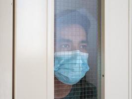 hombre solitario con mascarilla médica mirando por la ventana. aislamiento en casa para la autocuarentena. concepto de cuarentena domiciliaria, prevención covid-19. situación de brote de coronavirus foto