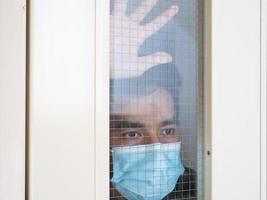 hombre solitario con mascarilla médica mirando por la ventana. aislamiento en casa para la autocuarentena. concepto de cuarentena domiciliaria, prevención covid-19. situación de brote de coronavirus foto