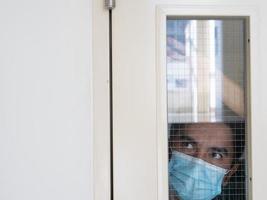 hombre solitario con mascarilla médica mirando por la ventana. aislamiento en casa para la autocuarentena. concepto de cuarentena domiciliaria, prevención covid-19. situación de brote de coronavirus foto