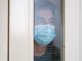 Lonely man in medical mask looking through the window. Isolation at home for self quarantine. Concept home quarantine, prevention COVID-19. Coronavirus outbreak situation photo