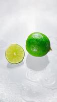 The green lime is blank with the cut lime slice showing the inside of the wet lemon pulp on a clear glass surface, reflecting the shadows of the lime and the wet water, giving it its freshness. photo