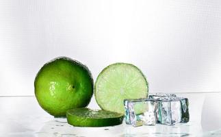 The green lemon is blank with a cut piece showing the inside of the citrus zest. The clear glass reflects the shadows of the lemon and the wet water, giving it freshness and ice adding cooling. photo