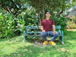 A young, visually impaired Asian man sits alone on a bench. in the city park