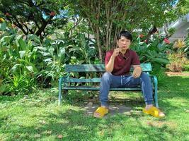 Young Asian man with black hair is sitting on a bench holding sunglasses. What are you thinking in the city park photo