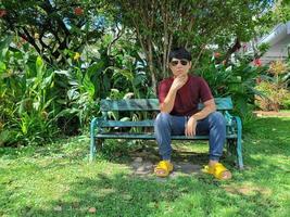 un joven asiático con el pelo negro está sentado en un banco. ansioso. ¿Qué estás pensando en el parque de la ciudad? foto