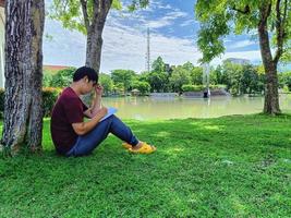 un joven asiático con gafas de pelo negro se sienta debajo de un árbol sosteniendo un libro. estrés y ansiedad. ¿Qué estás pensando en la ciudad del parque de verano? foto
